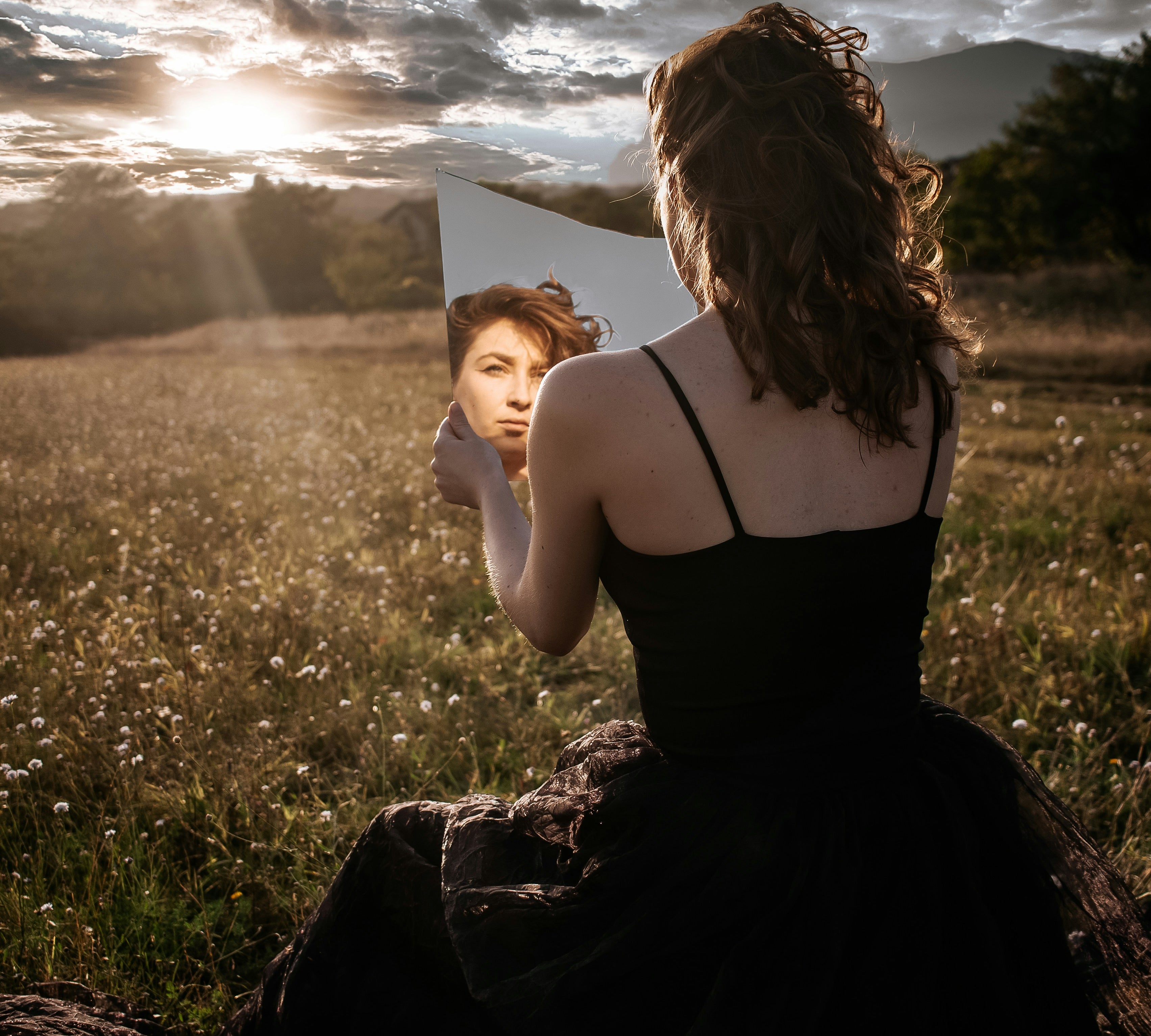 A woman gazes into a mirror while sitting in a serene field at sunset, symbolizing introspection, self-reflection, and the connection between inner beauty and nature. The image highlights themes of beauty, energy, and self-awareness.  