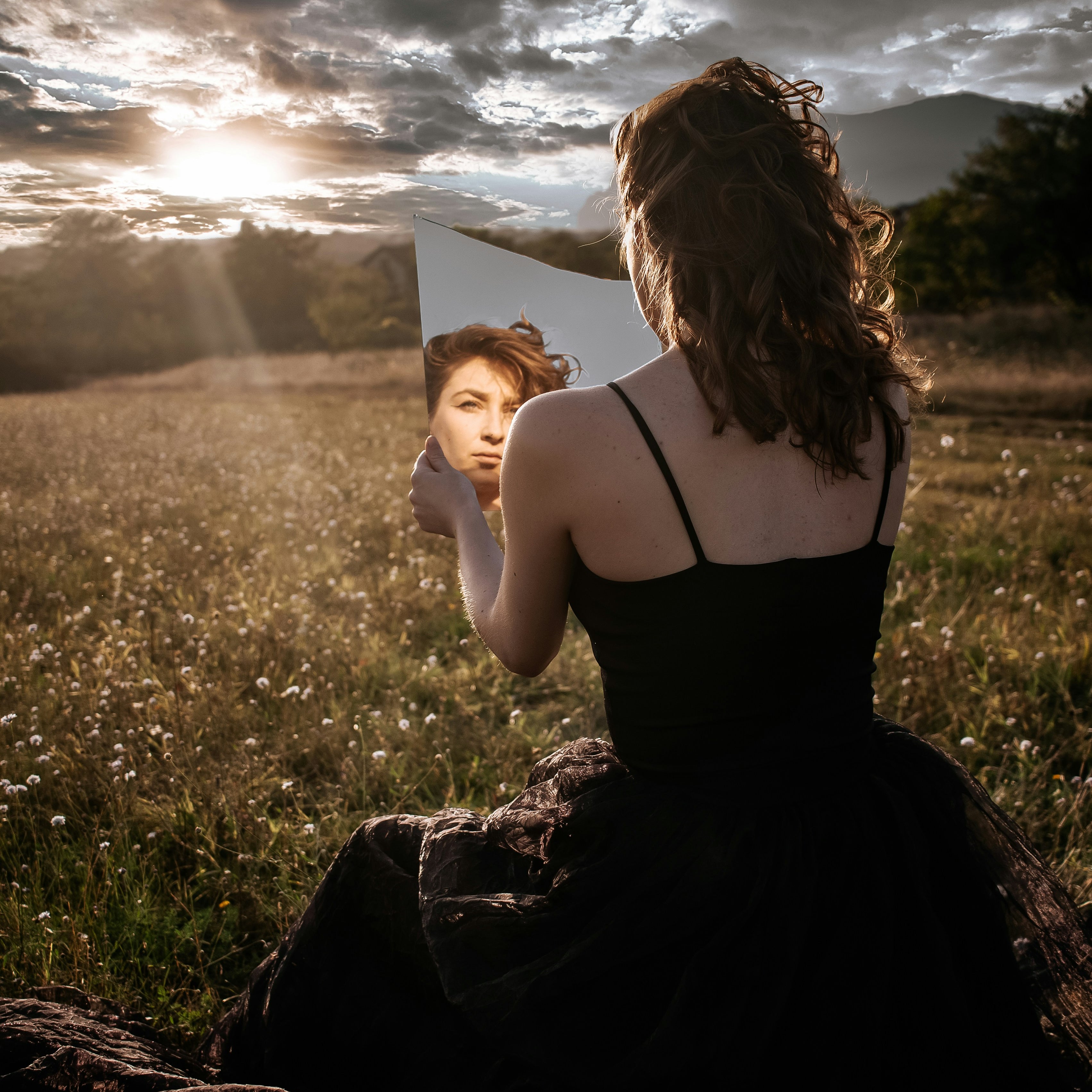 A woman gazes into a mirror while sitting in a serene field at sunset, symbolizing introspection, self-reflection, and the connection between inner beauty and nature. The image highlights themes of beauty, energy, and self-awareness.  