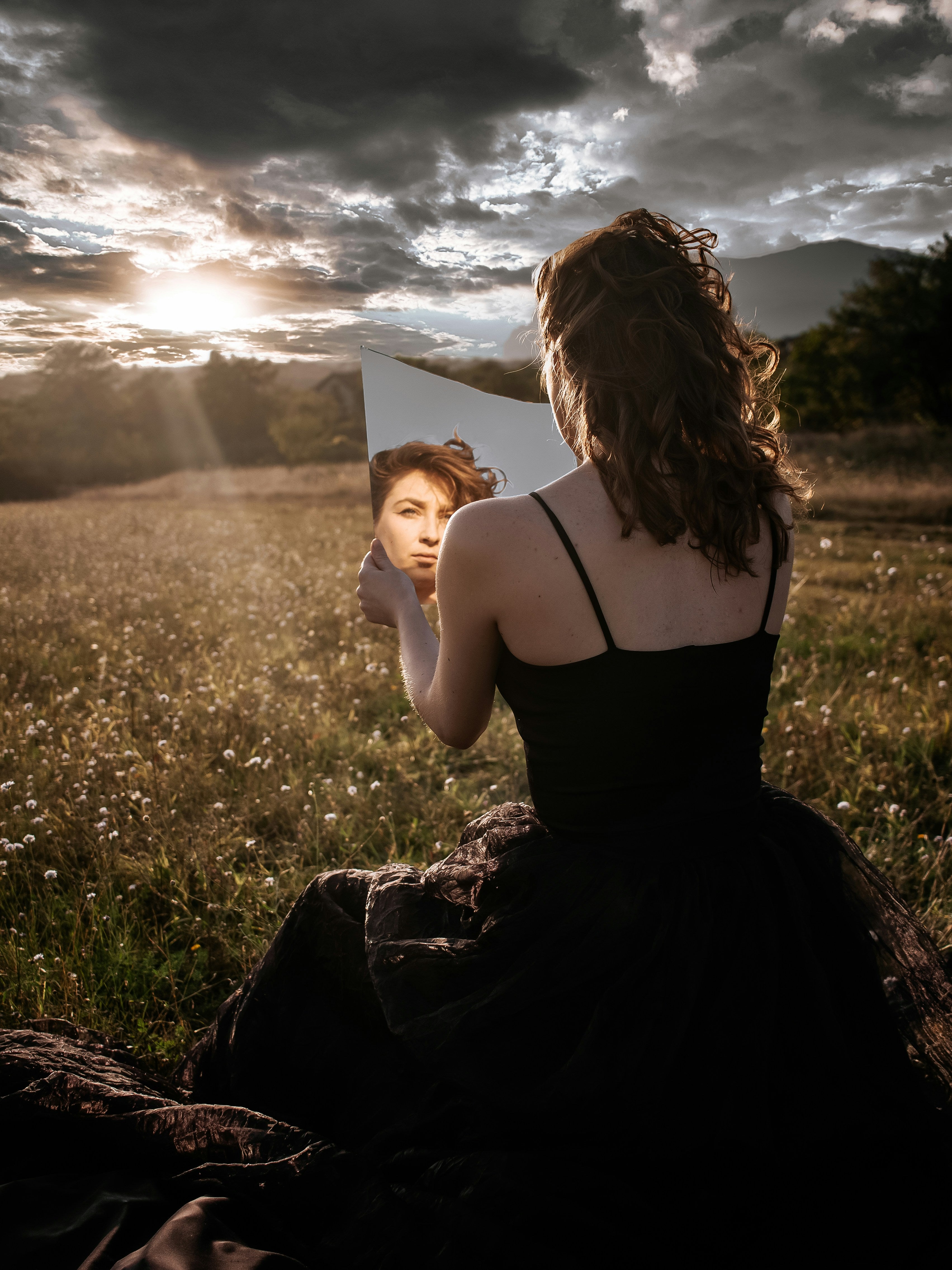 A woman gazes into a mirror while sitting in a serene field at sunset, symbolizing introspection, self-reflection, and the connection between inner beauty and nature. The image highlights themes of beauty, energy, and self-awareness.  