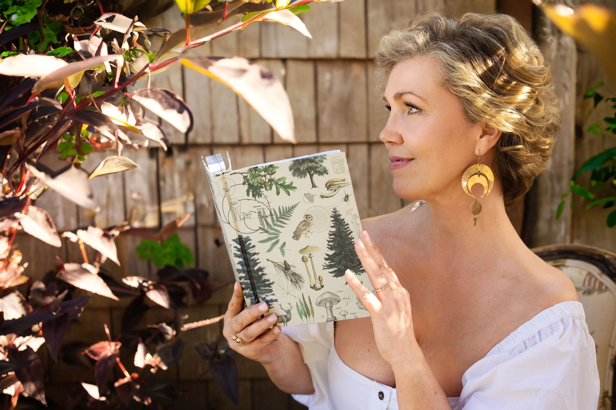 Amy Wall, founder of Higher Self Beauty, holding a nature-themed journal and looking contemplatively at the garden, symbolizing the connection between natural beauty and inner reflection.